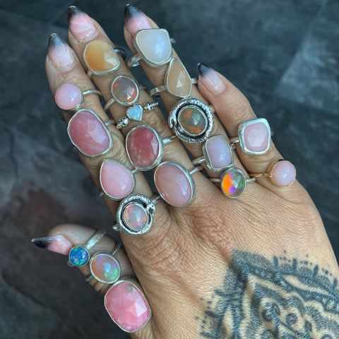 many different shapes of opal rings, pastels, fiery colours and white opals on a tattooed hand with a grey background