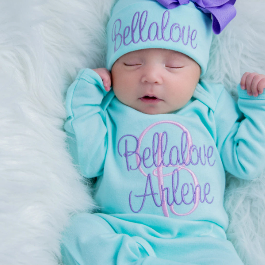 A baby girl wearing the Newborn Girl Personalized Aqua and Purple Hat & Romper Outfit from Junie Grace. 