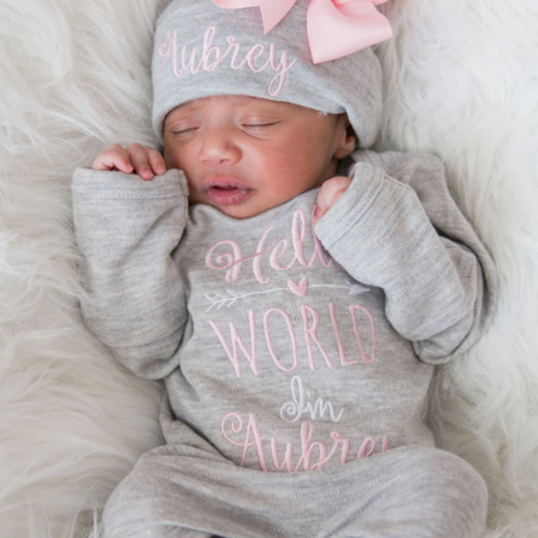 A baby girl wearing the “Hello World” Personalized Baby Girl Romper Set With Matching Bow Hat from Junie Grace. 