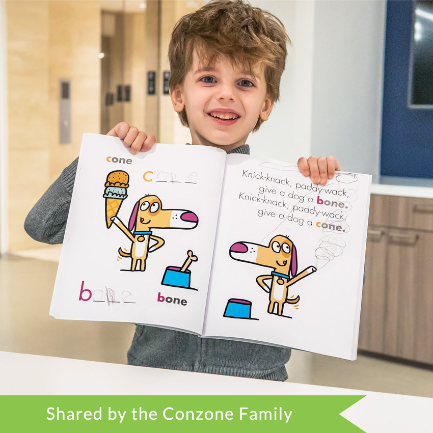 A customer photo of a smiling brunette boy holding up the I can Doodle Rhymes book. The left page shows an excited dog holding and looking up at an ice cream cone. Next to the dog is a bone in a blue bowl. The text on the page says "cone" and "bone." The right page shows a mirror image of the left page illustration, missing the bone and ice cream cone. The text reads "knick-knack, paddy-wack, give a dog a bone. Knick-knack, paddy-wack, give a dog a cone."