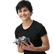A teenage child with short black hair is smiling and holding a completed Robotis Bioloid Stem project. The structure has black wheels in the back and the light gray pieces are flat on the bottom and is a large block in the middle on top with red and black wiring coming out from the block and into batteries.