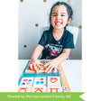 A customer photo of a black-haired girl sitting at a white table, smiling large with her teeth all showing and her eyes squinted from the smile. She has her hands placing a Bambino L U K tile on the control board. You can see shaped on the tiles that you match to the book under the see-through controller. There are gold spots on the white wall behind her.