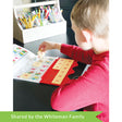 A customer photo of a young blonde boy sitting at a table and playing with the MiniLUK. He is holding tile # 3, getting ready to place it in the controller on the table. The controller is clear and red with peach-colored tiles with red numbers on them. The controller is set over one of the game books. You can see colored arrows pointed in different directions that you will match the tiles to.