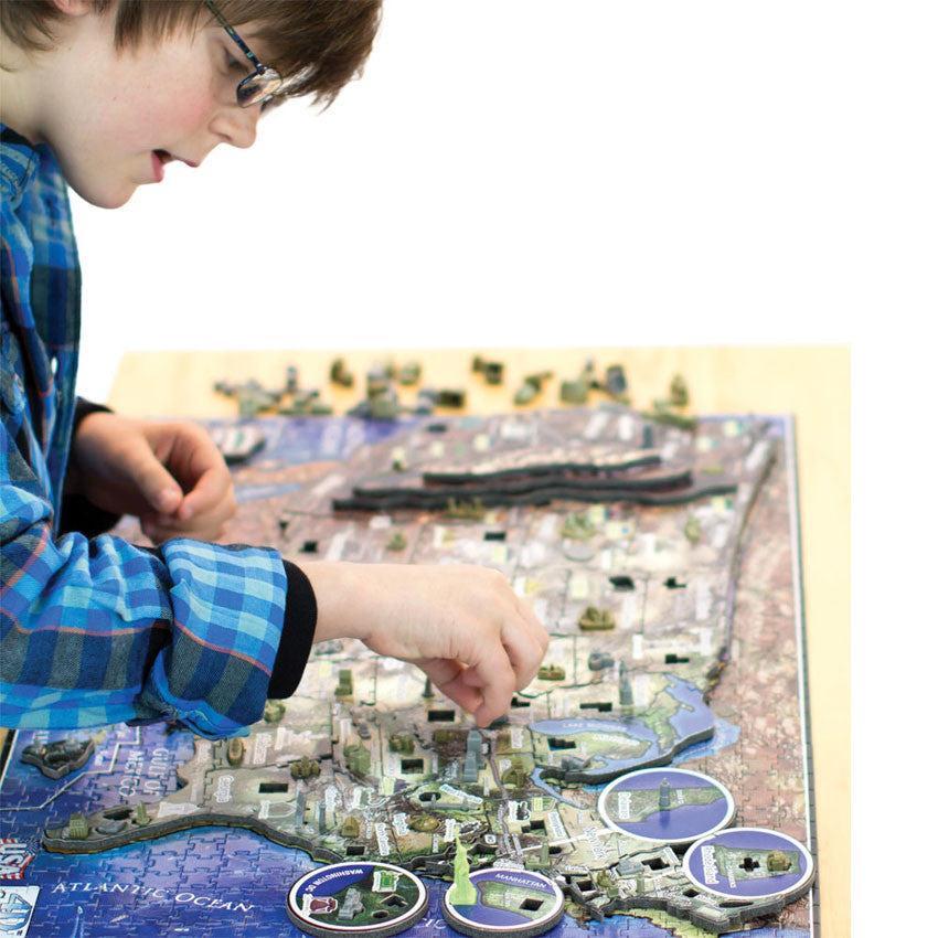 A young boy with dark hair and glasses is in the process of putting a piece into the U S A History Over Time Puzzle. The puzzle base is completed, showing the different levels as the USA is a taller level than the ocean base. There are cut-out’s to put in each states’ famous building. Out of focus in the background, you can see all the building pieces.