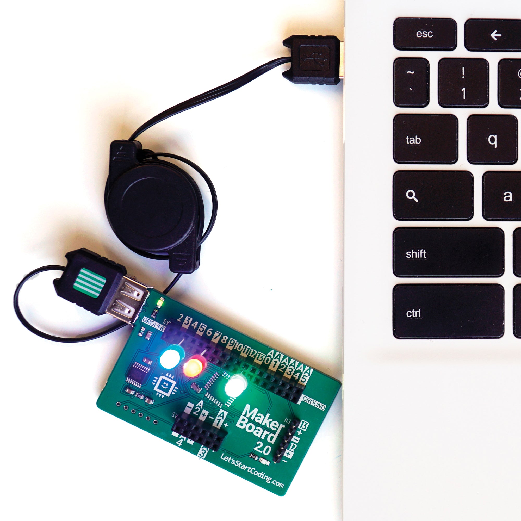 Let’s Start Coding Base Kit Maker Board plugged into a computer with the retractable U S B plug.  The Maker Board is dark green with black grid components for plugging in L E D's and buttons. There are 3 L E D lights plugged into the board and are lit up. You can see the left side of a laptop keyboard on the right.