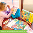 A customer photo of a dark-blonde girl painting with the Djeco Natural World Workshop. She has her elbows resting on the table in front of her, painting on the desert oasis project, which is resting on a blue, pillow-like easel. On the table, to her right is the product box and the rest of the contents laid out, including the paint tray that she is using. The colorful projects that are visible are the underwater scene on the box and the jungle scene, that is on top of the other project sheets.