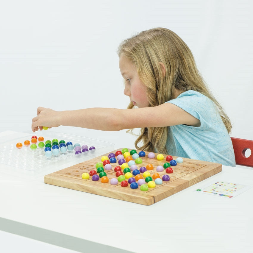 A blonde girl is in the middle of playing ColorKu. The board in front of her has many pieces in place on top. Off to the right side is a challenge card. The girl is leaning over with her left hand and is grabbing a yellow ball from the ball tray on her right. The tray has rows of balls waiting to be added to the board. The colored balls are red, orange, yellow, bright green, dark green, light blue, dark blue, light purple, and dark purple.