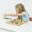 A blonde girl is in the middle of playing ColorKu. The board in front of her has many pieces in place on top. Off to the right side is a challenge card. The girl is leaning over with her left hand and is grabbing a yellow ball from the ball tray on her right. The tray has rows of balls waiting to be added to the board. The colored balls are red, orange, yellow, bright green, dark green, light blue, dark blue, light purple, and dark purple.