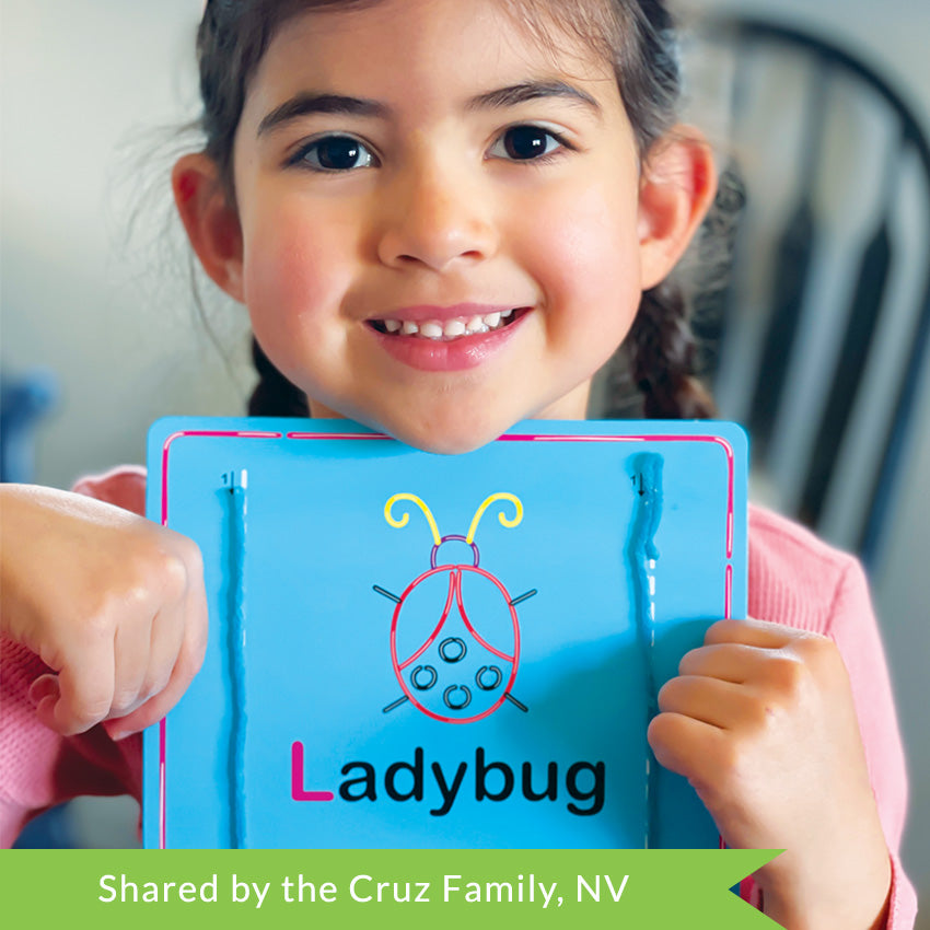 A customer photo of a young brunette girl smiling and holding up a Wikki Stix Alphabet card. The card is blue with a ladybug in the middle and an uppercase and lowercase L on it. She has used blue wax sticks to cover the letters on the board.