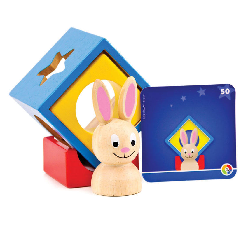 The Bunny Boo game setup with a challenge card. The wood blocks are stacked with the red piece that shows a half circle piece cut out of the middle on the bottom. Stacked on top and tilted to a diamond shape is a hollow blue wood block that has shapes cut out on the sides with a yellow piece with a hollow circle through the middle tucked inside the blue block. The bunny is sitting in font of the stacked blocks. The blue challenge card is propped up against the blocks, showing the shapes stacked as they are.