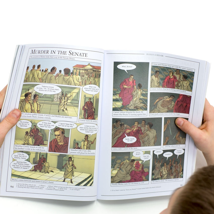 Graphic Shakespeare book, being held open by young hands, to show inside pages. The book is a comic book style layout with squared illustrations and talk bubbles. The pages are of a scene in Julius Caesar, titled “Murder in the Senate.” Senators gather in the Senate House and Brutus kisses Caesar’s hand which initiates more men to come up and attack Caesar and stab him to death.