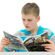 A young blonde boy in a striped shirt is reading the Graphic Shakespeare book. The cover is mainly black with white text and illustrations of the plays inside. The cover shows that the book includes; Romeo & Juliet, Julius Caesar, Hamlet, Macbeth, and A Midsummer Night’s Dream.