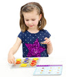 A young brunette girl with a sequins star shirt is playing the Smart Cookies game. She has a blue, square, piece in her left hand and is pointing to the top-right square on the game board with her right hand. The game board in front of her is filled with the red, yellow, and blue cookie pieces, except the one space she is pointing to. To her upper-left is the spiral-bound game challenge book.