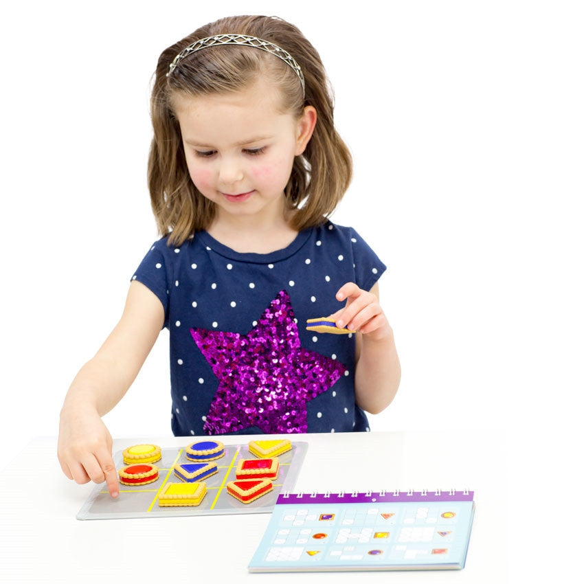 A young brunette girl with a sequins star shirt is playing the Smart Cookies game. She has a blue, square, piece in her left hand and is pointing to the top-right square on the game board with her right hand. The game board in front of her is filled with the red, yellow, and blue cookie pieces, except the one space she is pointing to. To her upper-left is the spiral-bound game challenge book.