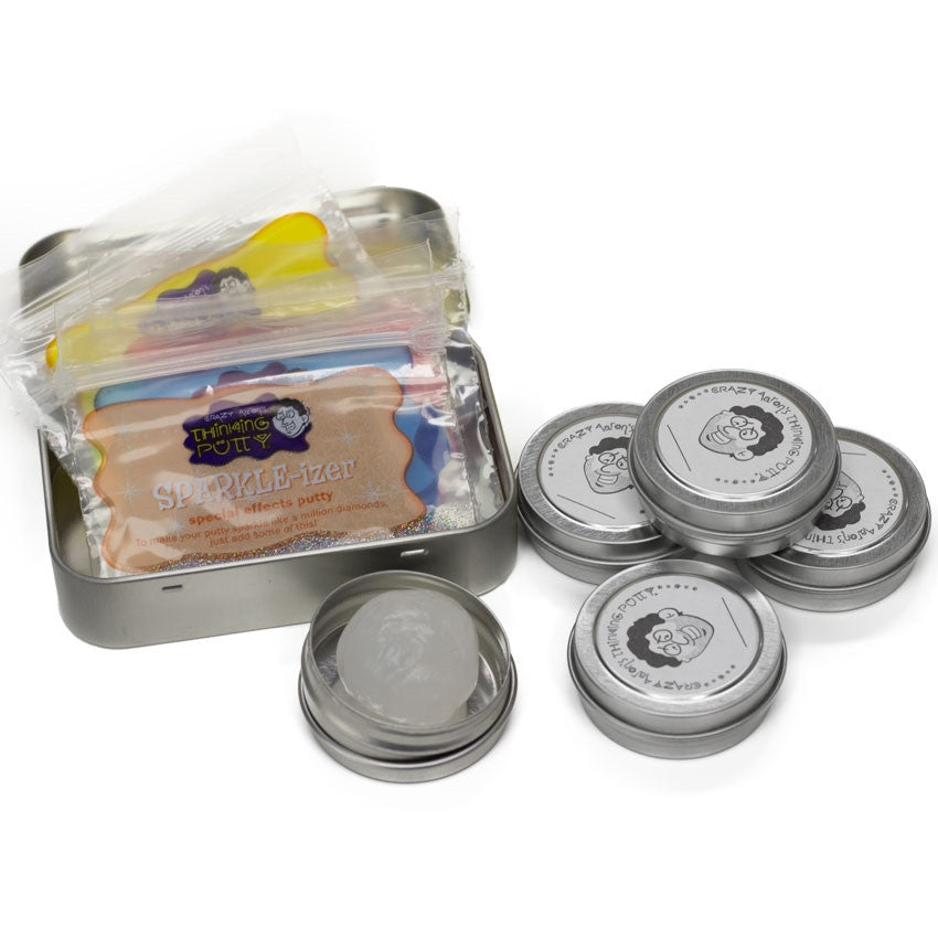 Some of the Mixed by Me, Glow, Thinking Putty Kit contents. A mint-style tin to the left shows several rectangular plastic bags containing condensed putty mixes. To the right is 5 tins of clear putty. One putty tin is open, showing a ball of the clear putty. The remaining 4 are closed with a label placed on the lids. The labels show a cartoon version of the business owner, Crazy Aaron.