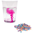 Color fizzers action shot of a glass with water in the process of being colored by a red tablet is in the upper-left. In the lower-right is a pile of many tablets colored blue, orange, and purple.