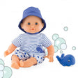 Corolle’s Bebe Marin. The light skinned baby doll has blue blinkable eyes and is sitting next to a rubber whale toy. He is wearing shorts, a shirt, and a blue bucket hat. His shirt is white with blue stripes and a whale patch on his lower-left. His shorts are white with a blue whale pattern. In the background are cartoon bubbles, randomly placed.