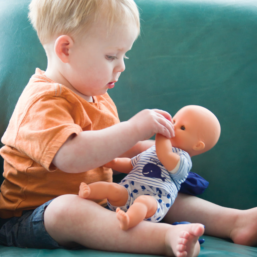 A young blonde boy sitting on a couch and holding Corolle’s Bebe Marin on his lap. He is holding baby Marin’s left arm up to his face as if he is trying to help him feed himself. The light skinned baby doll has blue blinkable eyes and is wearing white whale shorts and a striped shirt with a whale patch. You can barely see the baby’s hat and whale toy laid on the couch and the boys leg.