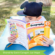 A customer photo of a young dark-haired boy wearing a baseball cap and laying on a yoga mat on the grass and holding the Sharpeners Geography Grade 2 book. The book is open toward the boy and shows the cover to the camera. The book cover is mainly orange with a white top containing the title and 2 sample pages in the middle. To the right is a huge pencil illustration, standing from bottom to top. The back cover is white with images of several books from the publisher.