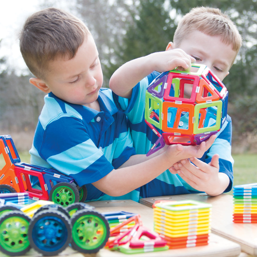 2 blonde boys in striped shirts are outside and are putting together a large shape, made up of the GeoSmart Educational Deluxe pieces on a picnic table. The pieces are geometrical shapes in squares and triangles and are a variety of different colors.  The boy on the left is holding up a large rounded object in the air from the bottom, while the boy on the right is holding it up on the top and the bottom. There are pieces in stacks and piles on the table, along with a car made from the pieces on the left.