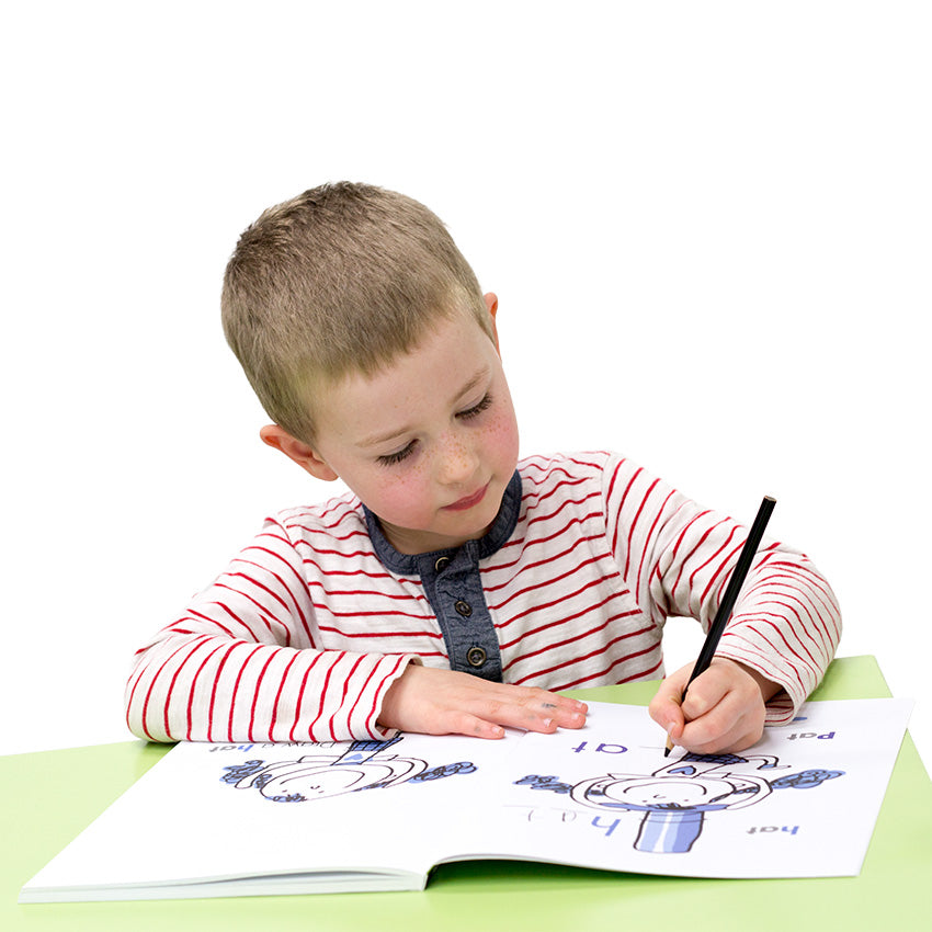 A dark blonde, freckled, boy in a striped shirt is sitting at a green table and writing with a black colored pencil in the I Can Doodle Rhymes book. The left book page shows an illustration of a girl in a hat with the word "hat" on the page. On the right page is a an illustration of a girl missing a hat with the instructions "draw a hat."