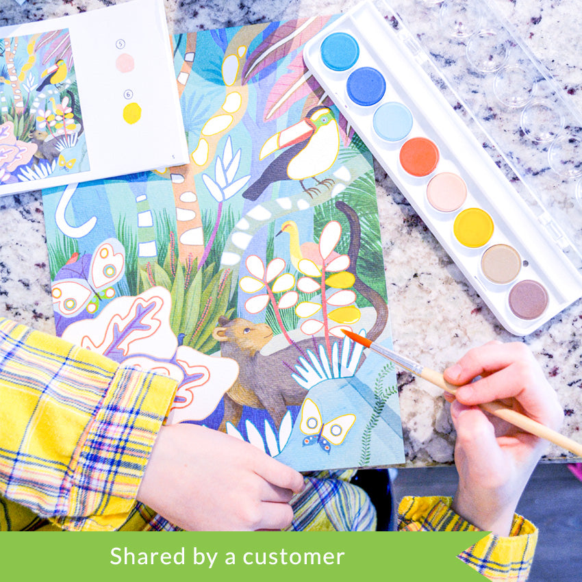 A customer photo of hands resting over the Djeco Natural World Workshop jungle project that are sitting on top of a marble table. In the top-left is the open instruction book. In the top-right is the paint tray. In the bottom-right, the hand is holding the paintbrush over the project page.