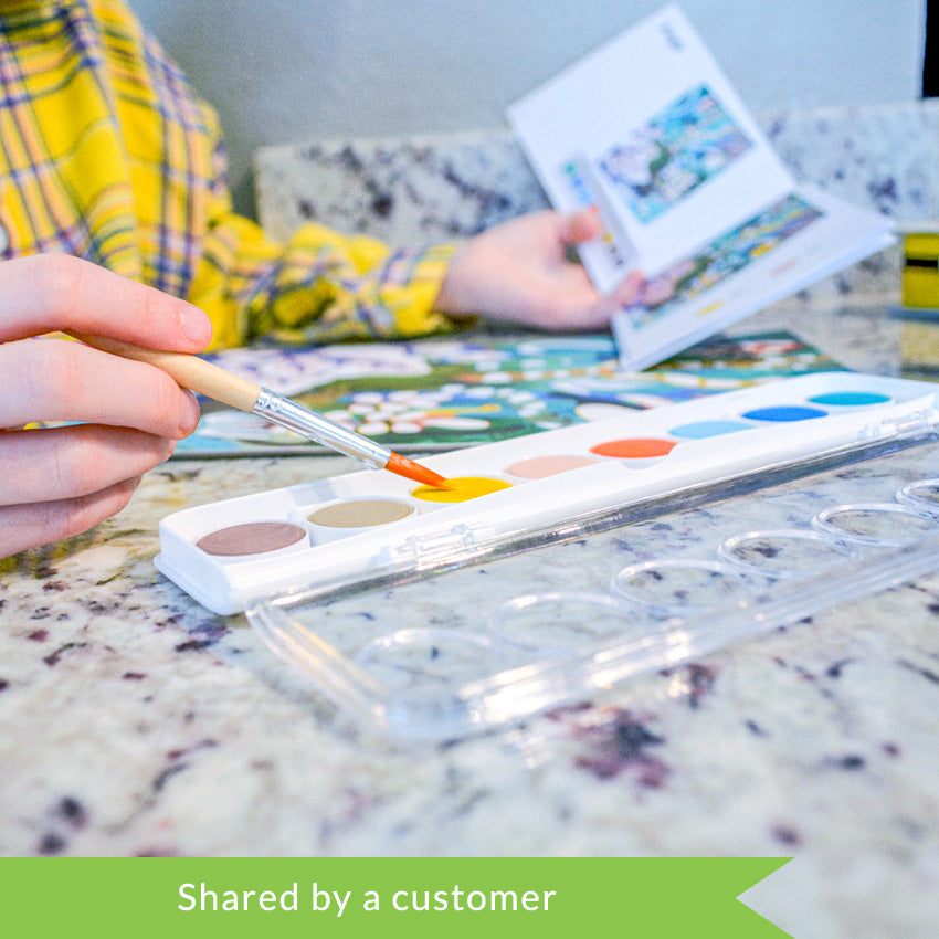 A customer photo of a child’s hands in the middle of working on a Djeco Natural World Workshop project. On top of a marble surface is a paint tray, project page, and instruction book. The child is holding the instruction book open with her left hand and holding the paintbrush with her right, dipping it into the yellow paint.