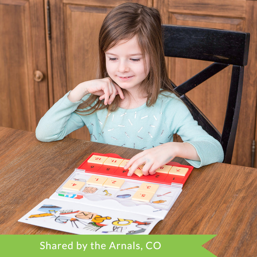 A customer photo of a young brunette girl sitting at a table and playing with the MiniLUK. She is holding tile # 6, getting ready to place it in the controller on the table. The controller is clear and red with peach-colored tiles with red numbers on them. The controller is set over one of the game books. You can see close up’s of objects on the top page, and the objects that match on the bottom page. The controller is placed over the book, so you can put the tiles down to match.