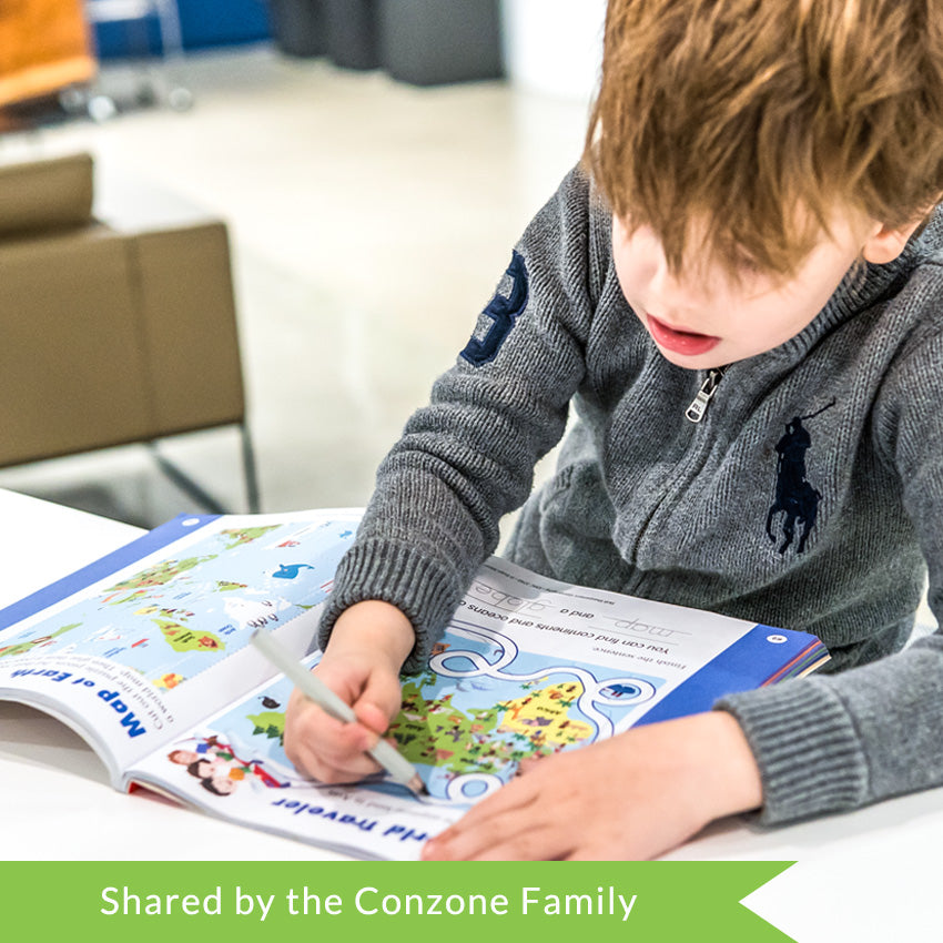 A customer photo of a young brunette boy drawing on a path in the Skill Sharpeners Geography Grade K book. The white path, the child is drawing on, is looped around a map of Europe, Asia, and Africa. On the right page is a map of the world. The pages are white with a blue border on the outer edges. The maps on the pages are colorful.