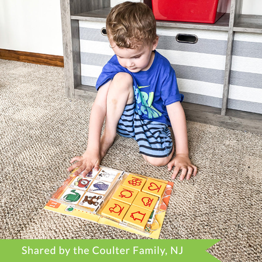 A customer photo of a young blonde boy sitting on the carpet and placing his hand on the Bambino L U K controller that is on top of a game book. You can see the book through the controller and shapes on the book and tile, that you match up. The yellow tiles have a duck, apple, flower, heart, car, and house on them. The book under shows a watering can, clock, candy, teddy bear, teapot, and snowman.