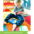 A customer photo of a blonde toddler sitting on the floor and holding a What’s Going on Here Conversation Card. The card is red and is large in his hands. Sitting between his legs is the card box. Toward the bottom of the picture, slightly under his foot, are more cards, of many colors, fanned out on the colorful square carpet.