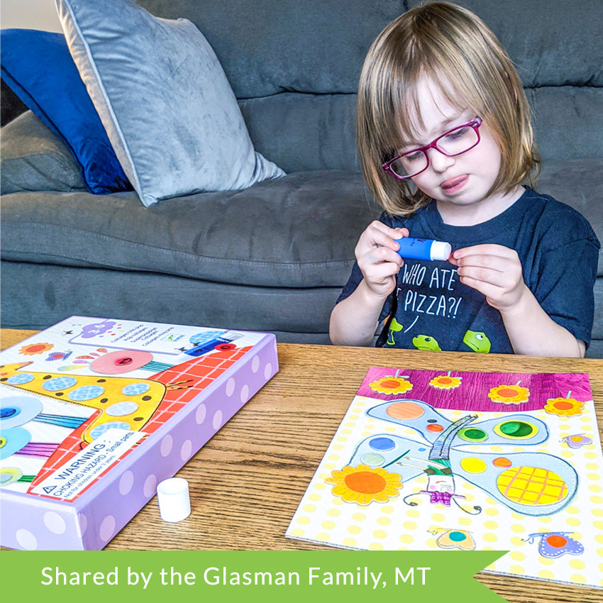 A customer photo of a blonde girl sitting in front of a coffee table working on a Djeco So Pop Collages project. She is looking down at the dot she is holing in her hand and a glue stick in the other with her tongue out as she is concentrating. The project in front of her is a butterfly with flowers. There are many dots and shaped already glued onto the page. To the left is the project box with a giraffe on the cover.