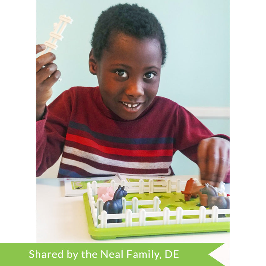 A customer photo of a black-haired boy holding a fence piece to the Smart Farmer game in his right hand and in in the process of grabbing an animal piece with his left hand. In front of him is the green Smart Farmer game board with white fence pieces all around the edge and farm animals inside, including; 2 pigs, 2 horses, and 2 cows. The instruction book is open in front of the boy to shows challenges.