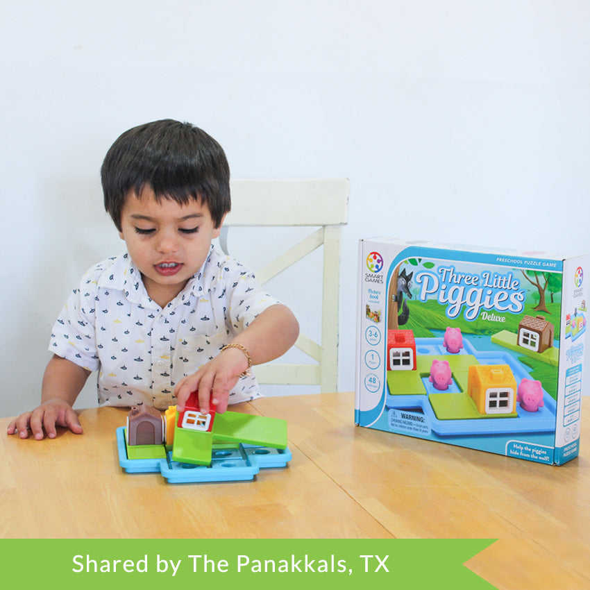 A customer photo of a young, dark-haired boy, playing with the Three Little Piggies game. He is sitting at a kitchen table and placing a red house piece on the blue game board in front of him. The game board is light blue with playing pieces on the board. The game box is standing on the table to the left of the boy.