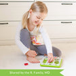 A customer photo of a smiling young blonde girl sitting on the floor and playing with the Smart Farmer game. She is holding a horse piece with her right hand and grabbing onto a sheep piece with her left hand. The game board is green and rectangular with white fence pieces all around the outside. Inside are a few more fence pieces with pigs and sheep. There is another horse and a feeding dish off to the side.