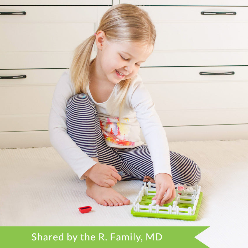 A customer photo of a smiling young blonde girl sitting on the floor and playing with the Smart Farmer game. She is holding a horse piece with her right hand and grabbing onto a sheep piece with her left hand. The game board is green and rectangular with white fence pieces all around the outside. Inside are a few more fence pieces with pigs and sheep. There is another horse and a feeding dish off to the side.