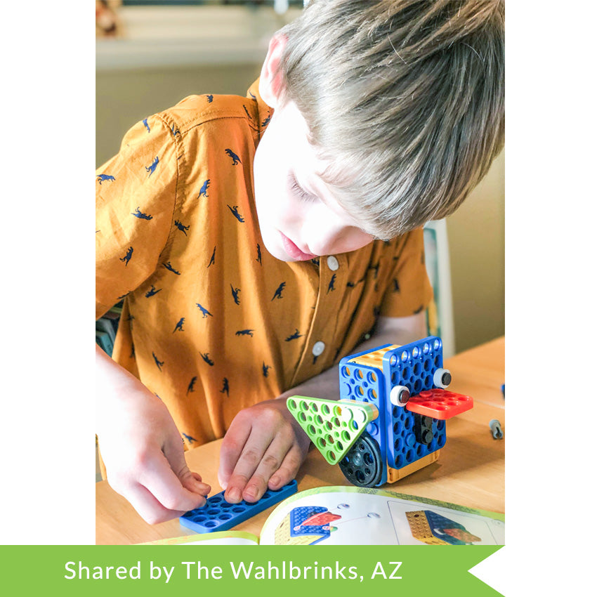 A customer photo of a blonde boy in an orange shirt with dinosaurs putting together a Robotis Play 600 Pets bluebird. On the table in front of him is the bluebird robot and the instructions laid out while he puts a peg in a blue piece.