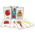 My First Touch and Feel Cards: First Words card pack open with 6 cards surrounding. In the middle is the blue packaging with a red apple. A card with a baby’s face can be seen inside the package. Surrounding the main pack are 6 cards, including; an apple, a teddy bear, jelly on toast, keys, a mirror, and corn. All items have spots for sensory texture and appearance. All cards have a white background with colored triangle-shaped flags all around the border.