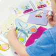 A close up of a child working on a Djeco So Pop Collages project. The child is gluing a dot to the large butterfly on the page. There are other dots previously glued onto the butterfly’s wings as well. You can see other cut out pieces on the table. In the upper-right you can see a lavender envelope with a sticker of the project on it. On top of that is the instruction book, opened to show steps of the butterfly project.