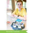 A customer photo of a young teenage boy sitting at a table playing with the Walls & Warriors game. He has the instruction booklet open with the game in front of him. The game base looks like a gray rock island with castle wall pieces and silver knight pieces with red and blue flags and shields placed on top. There are 3 red knight pieces off to the side.