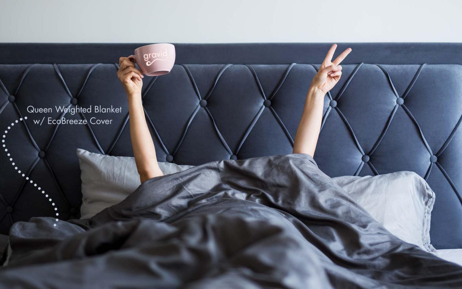 Person unwinding in bed with a cup, making a peace sign under a weighted blanket