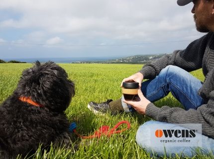 Man holding coffee in a KeepCup reusable cup next to black dog sitting in grass