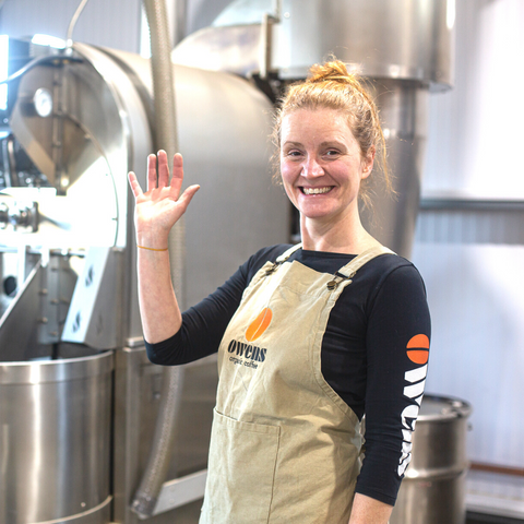 Woman in Owens Coffee apron smiling and waving with coffee roaster behind her