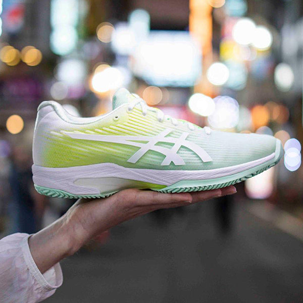 Image of a woman holding a white, green and yellow ASICS tennis shoe.