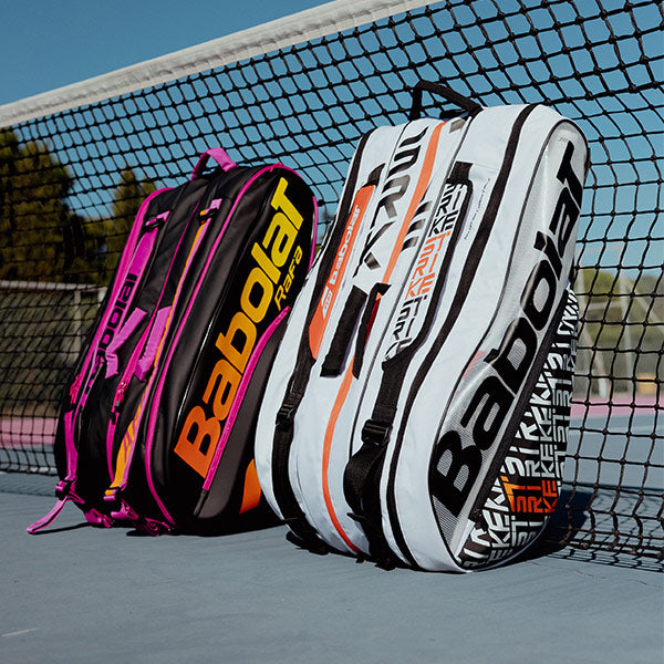 Shot of Babolat Babolat Pure Aero Rafa and Pure Strike 12 Racquet tennis bags leaning upright against a tennis net.