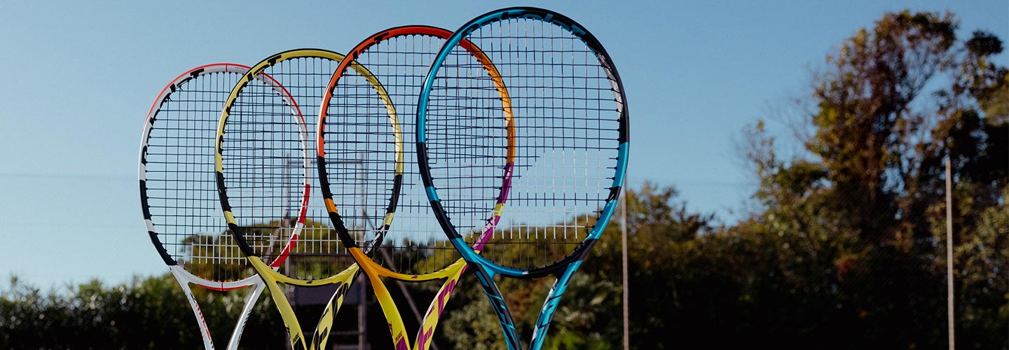 Babolat Pure Strike, Pure Aero, Pure Aero Rafa, and Pure Drive tennis racquets standing on an outdoor tennis court against a blue sky and green trees.