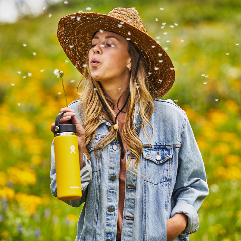 Woman outdoors blowing on dandelion while holding yellow Hydro Flask bottle