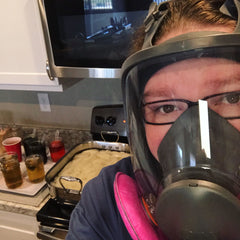 Image of me dyeing yarn in my kitchen wearing a full face respirator with dye and yarn behind me.