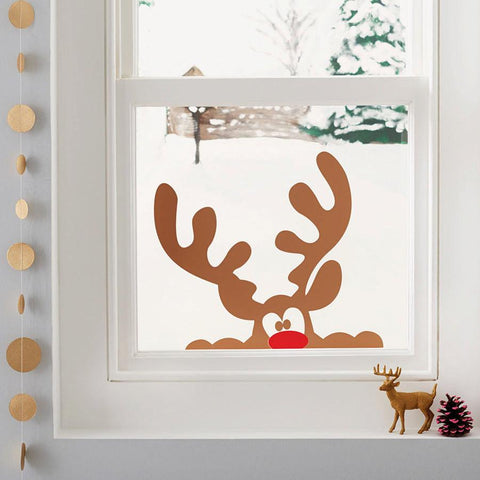 A Christmas window sticker of a cheeky brown reindeer peeping into the room, with his red nose just showing over the bottom of the window frame. It's snowing outside and there are other Christmas decorations around the window. 