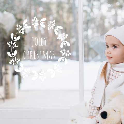 Girl standing next to a window with a white wreath-shaped window sticker with 'Jolly Christmas' written on it, in front of a snowy forest scene. 